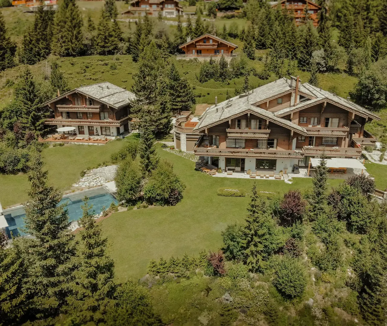Aerial view of luxurious alpine chalets surrounded by lush greenery and pine trees, featuring manicured lawns and a swimming pool under clear summer skies.