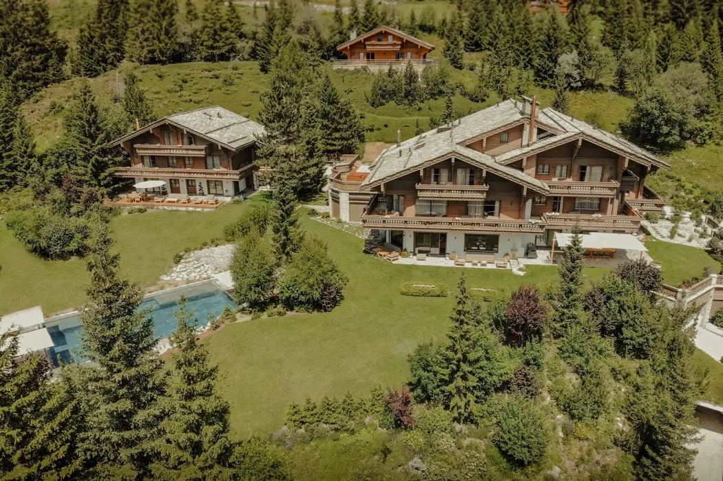 Вид с воздуха на роскошные альпийские шале, окружённые густой зеленью и соснами, с ухоженными газонами и бассейном под ясным летним небом.