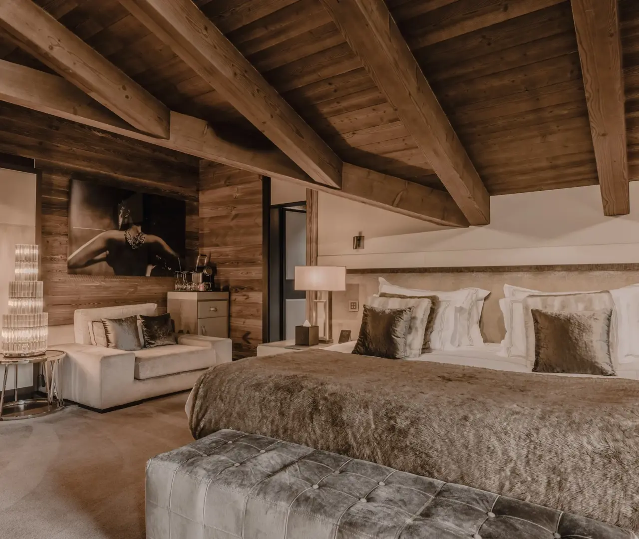 A lavish chalet bedroom with rustic wooden beams and walls, a large bed dressed in luxurious linens and fur textures, and soft ambient lighting. A plush sofa sits beside a crystal floor lamp and wall art, creating a refined yet cozy mountain retreat atmosphere.