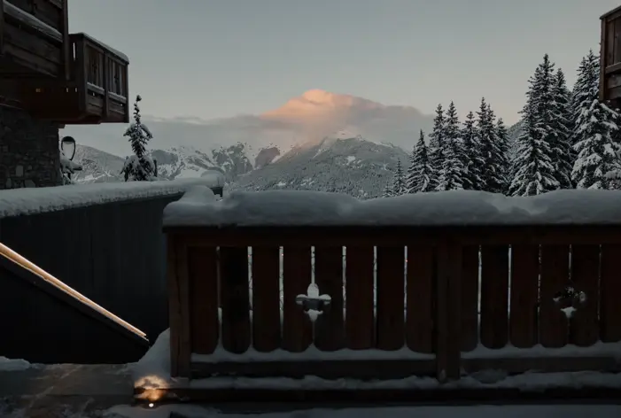 Paysage de montagne enneigée vu depuis le balcon d’un chalet en bois au lever du soleil, avec des sapins et des nuages roses au-dessus des sommets.