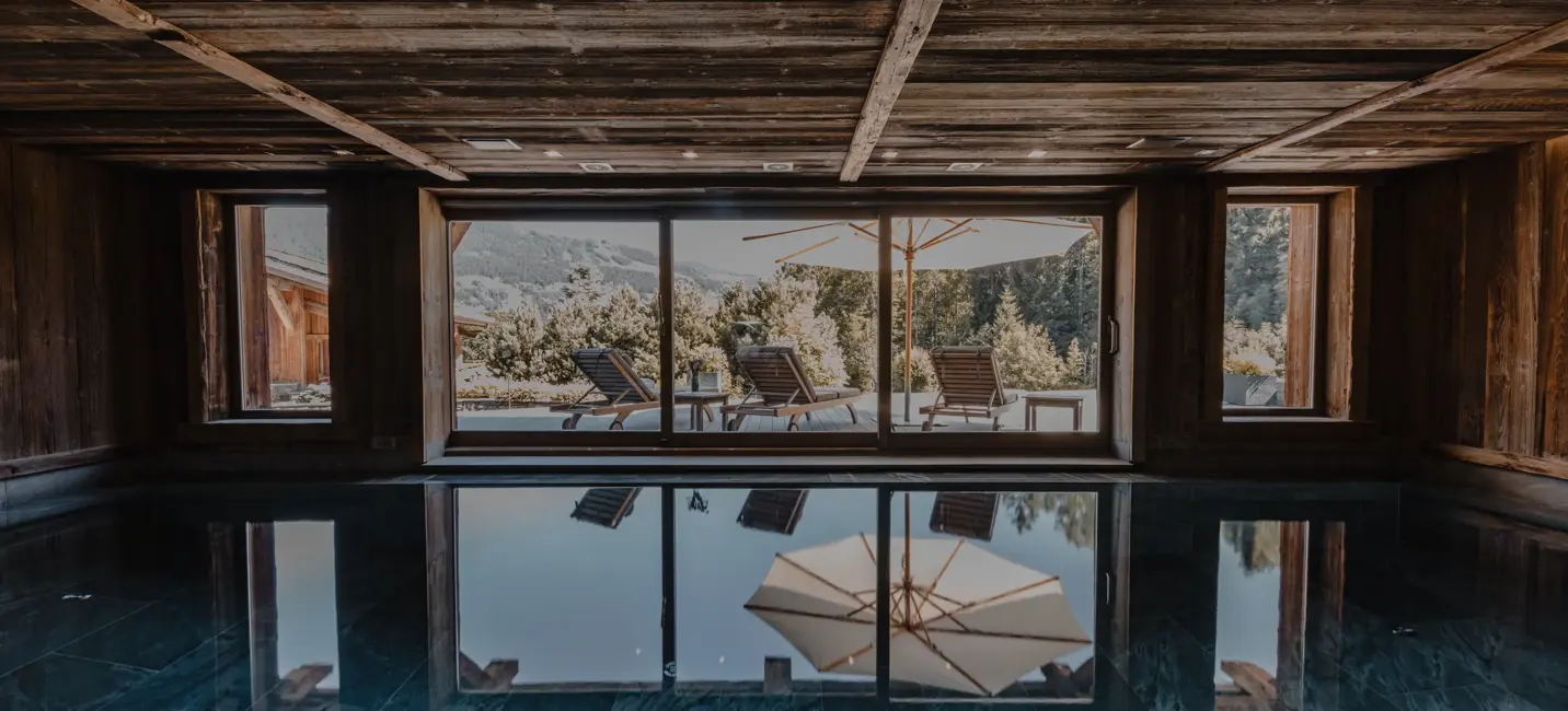 Indoor pool with a wooden ceiling reflecting the serene outdoor view through large glass doors, showing sun loungers and umbrellas on a terrace surrounded by mountains and trees.