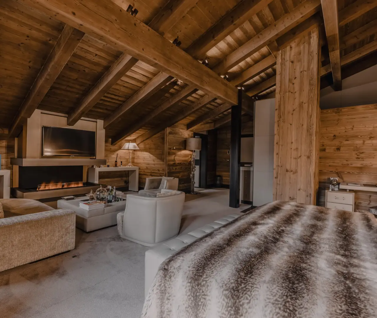 A warm, elegant suite under timbered ceilings, featuring a cozy lounge, modern fireplace, and plush bed. The natural wood tones and soft lighting create an inviting alpine charm.
