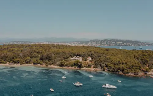 Aerial view of a lush Mediterranean island surrounded by turquoise waters, with yachts and boats anchored along the coastline and a distant cityscape under a clear blue sky.