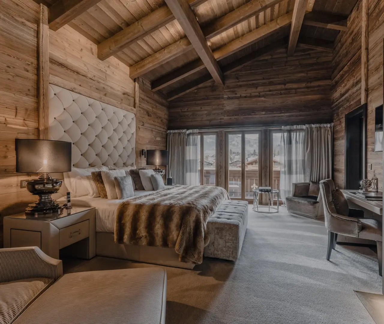 Luxurious chalet bedroom with warm wooden walls and vaulted ceiling beams, featuring a plush bed with a tufted headboard, fur throw, and elegant decor that opens to a balcony with mountain views.
