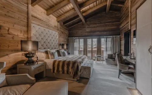 Luxurious chalet bedroom with warm wooden walls and vaulted ceiling beams, featuring a plush bed with a tufted headboard, fur throw, and elegant decor that opens to a balcony with mountain views.