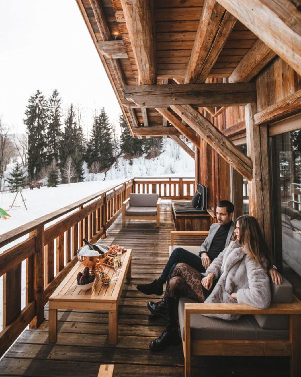 Couple relaxing on a wooden chalet terrace overlooking snowy mountains with food and wine on the table.