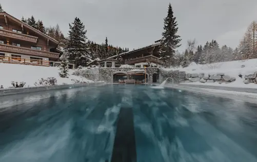 Piscine extérieure chauffée d’un chalet alpin de luxe entouré de pins enneigés, avec une architecture en bois chaleureuse et de la vapeur s’élevant des eaux turquoise dans un cadre de montagne paisible.