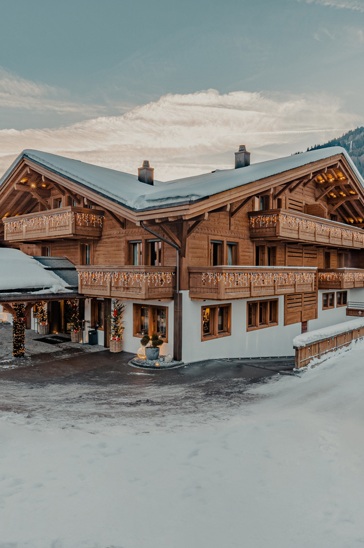Charming wooden ski chalet decorated with lights in a snowy alpine village, surrounded by mountains and winter scenery at sunset.