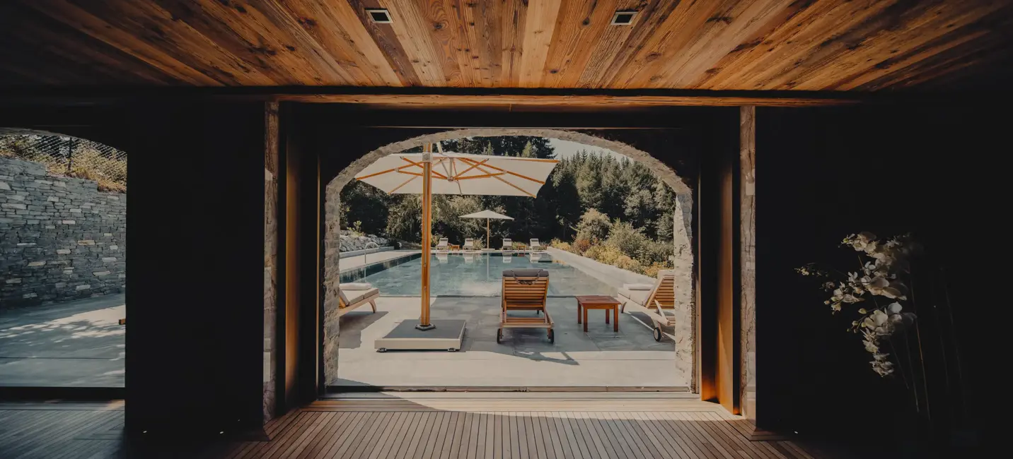 View from a wooden interior leading to an outdoor pool area with sun loungers, umbrellas, and lush greenery in the background.