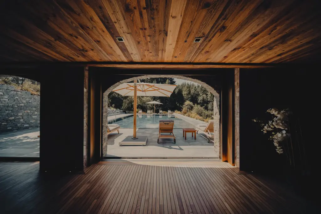 Vue depuis un intérieur en bois donnant sur une piscine extérieure avec des transats, des parasols et une végétation luxuriante en arrière-plan.