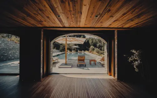 View from a wooden interior leading to an outdoor pool area with sun loungers, umbrellas, and lush greenery in the background.