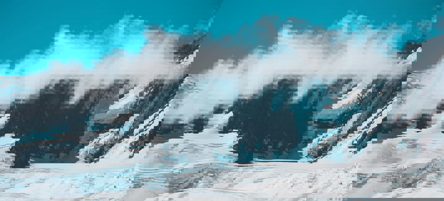 Un hélicoptère posé sur une vaste étendue enneigée, au pied de sommets glacés entourés de nuages et d’un ciel bleu limpide.