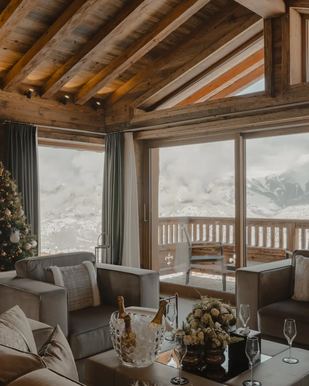 Salon de chalet de luxe avec vue panoramique sur la montagne, poutres en bois, sapin de Noël et champagne sur la table basse.