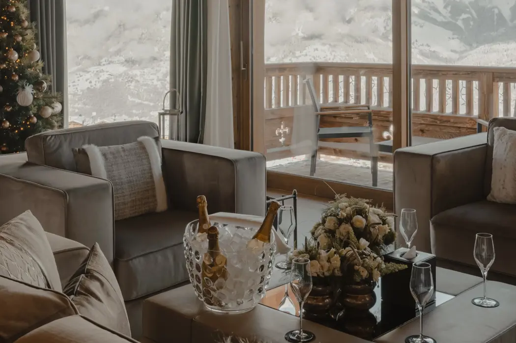 Luxury chalet living room with panoramic mountain view, wooden beams, Christmas tree, and champagne setup on the coffee table.