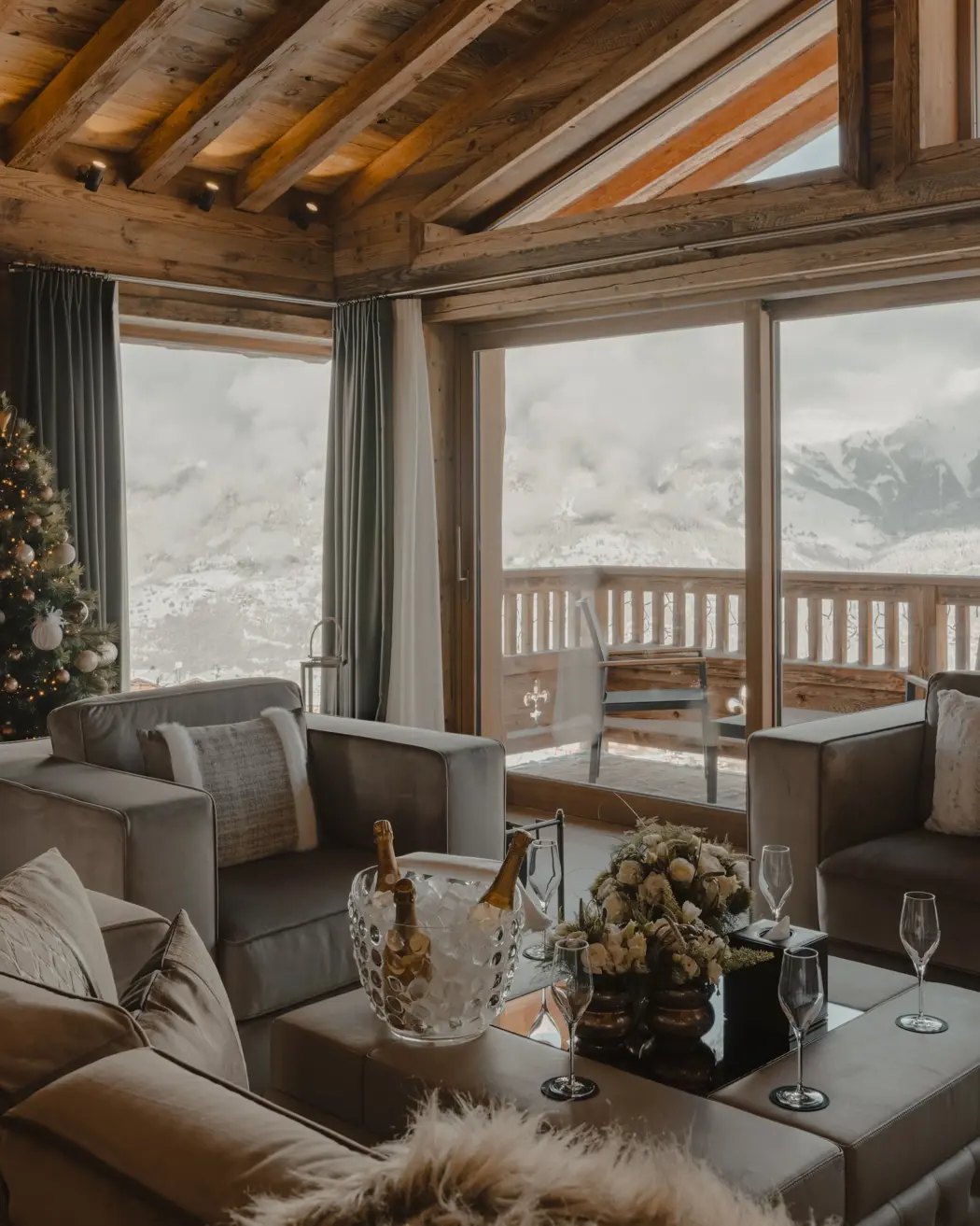 Luxury chalet living room with panoramic mountain view, wooden beams, Christmas tree, and champagne setup on the coffee table.