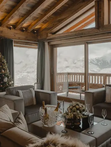 Luxury chalet living room with panoramic mountain view, wooden beams, Christmas tree, and champagne setup on the coffee table.