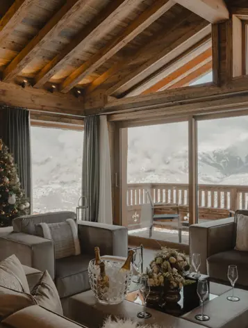 Luxury chalet living room with panoramic mountain view, wooden beams, Christmas tree, and champagne setup on the coffee table.