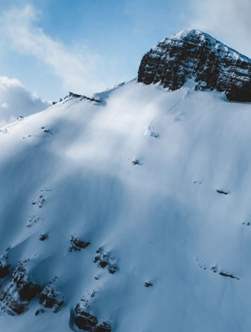 Snow-covered mountain peak with steep slopes and rocky cliffs under a blue sky.