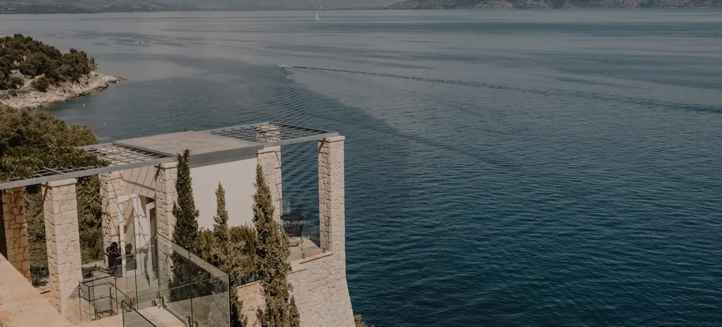 Villa élégante en pierre perchée sur une falaise côtière, avec terrasses en verre surplombant la mer bleue paisible et les montagnes au loin.