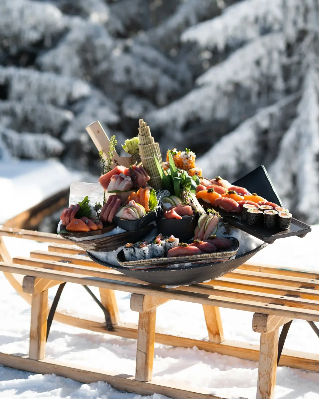 An artfully arranged sushi platter featuring fresh sashimi and rolls, displayed outdoors on a wooden sled against a snowy alpine backdrop.