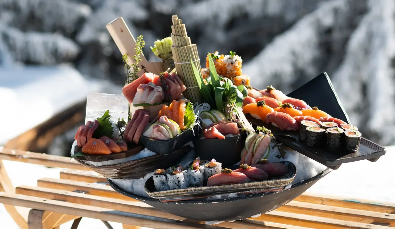 An artfully arranged sushi platter featuring fresh sashimi and rolls, displayed outdoors on a wooden sled against a snowy alpine backdrop.