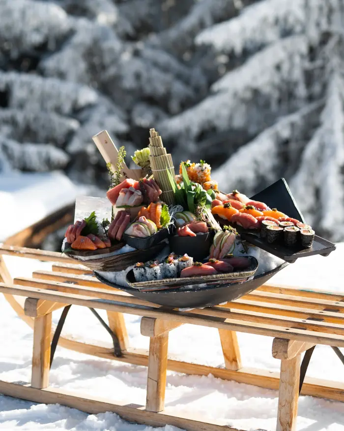 An artfully arranged sushi platter featuring fresh sashimi and rolls, displayed outdoors on a wooden sled against a snowy alpine backdrop.
