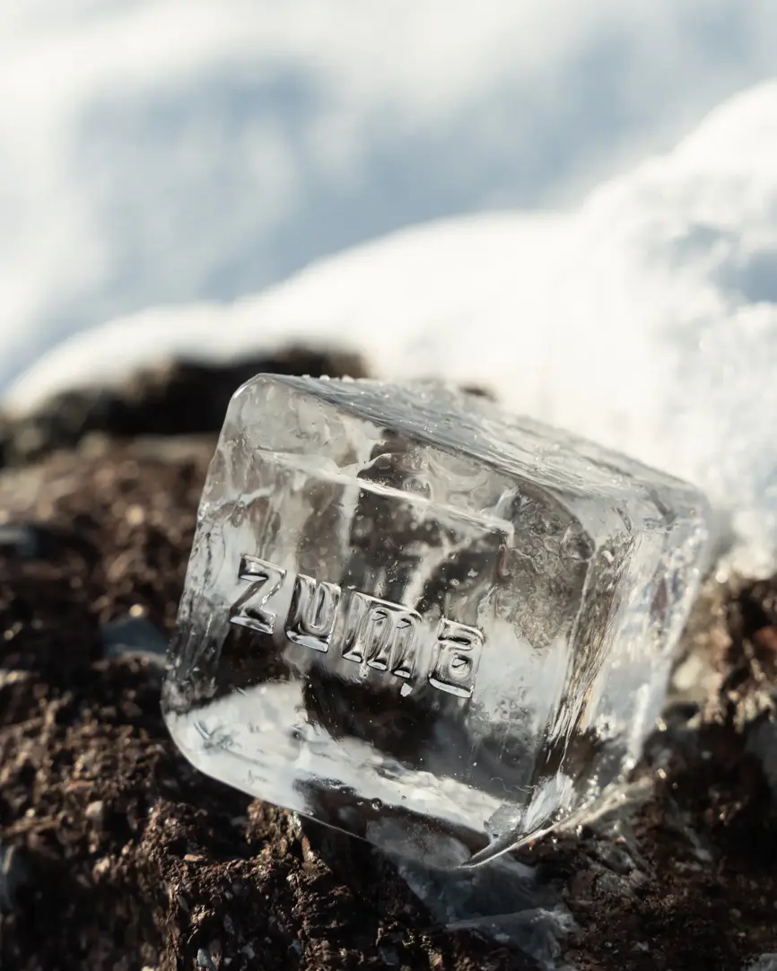 Clear ice cube with “Zuma” engraving resting on snow and rock.
