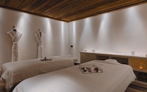 Luxurious spa room with two massage beds, soft lighting, wooden ceiling, and robes hanging on the wall.
