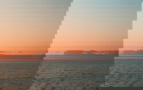 Tranquil seascape at sunrise with soft orange sky and distant mountain silhouette.