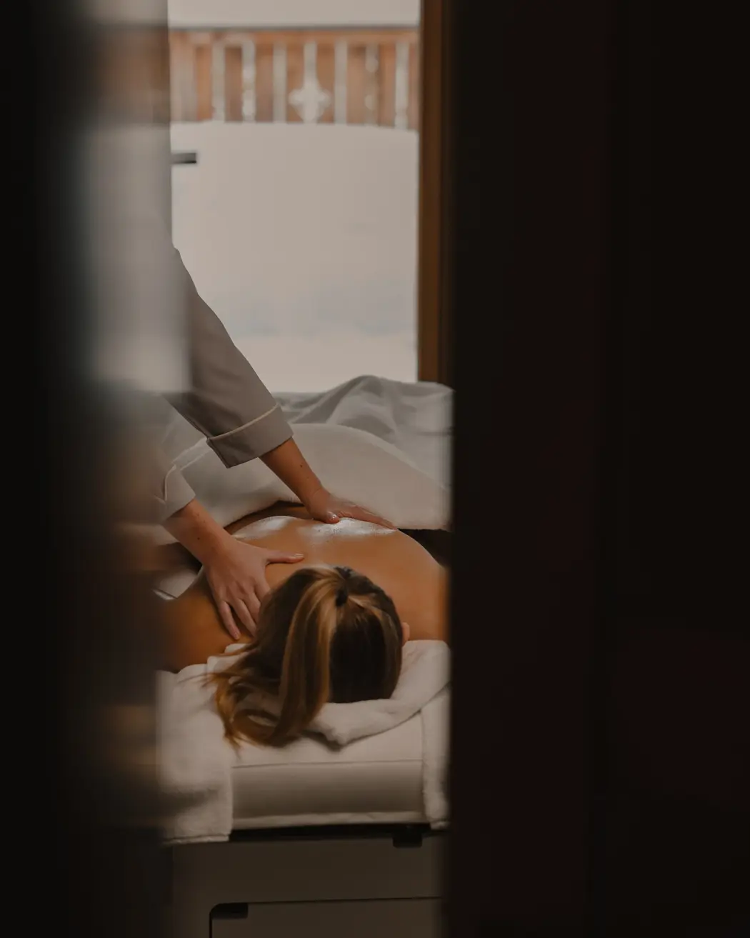 Personne recevant un massage du dos relaxant à l’intérieur, avec un éclairage doux et une vue sur un balcon en bois enneigé.