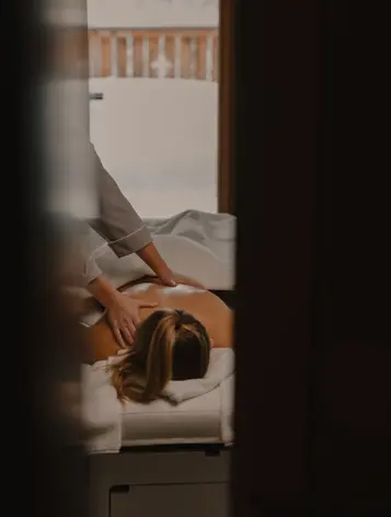 Personne recevant un massage du dos relaxant à l’intérieur, avec un éclairage doux et une vue sur un balcon en bois enneigé.