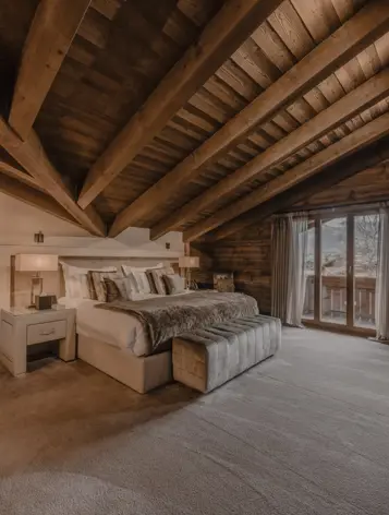 A luxurious chalet bedroom with exposed wooden beams, soft carpet flooring, and a large bed adorned with plush pillows and a fur throw. Warm lighting highlights the rich wood textures, while glass doors open to a balcony with mountain views, creating a serene alpine retreat.