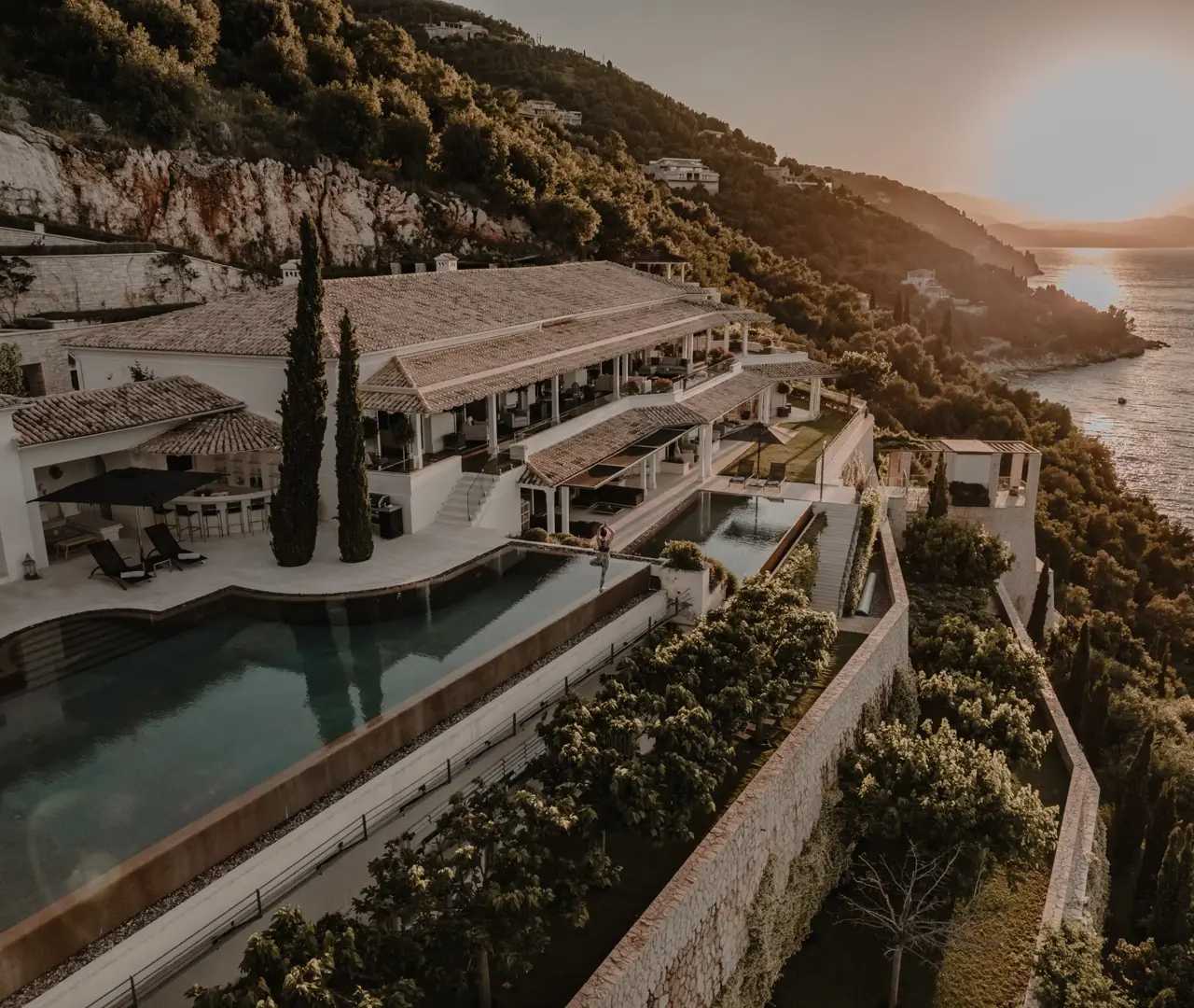 Aerial view of a luxury seaside villa built on a hillside, with terraces, pools, and a sunset over the ocean.