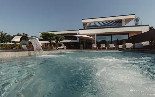 Современная роскошная вилла с инфинити-бассейном, водопадами, шезлонгами и стеклянной архитектурой под ясным голубым небом.