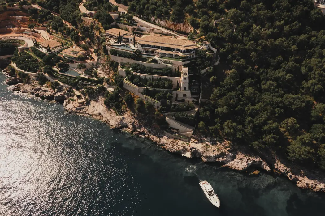 Vue aérienne d’un domaine luxueux à flanc de falaise, à l’architecture en pierre en terrasses, entouré d’une végétation luxuriante et surplombant la mer bleue scintillante où dérive un yacht blanc.