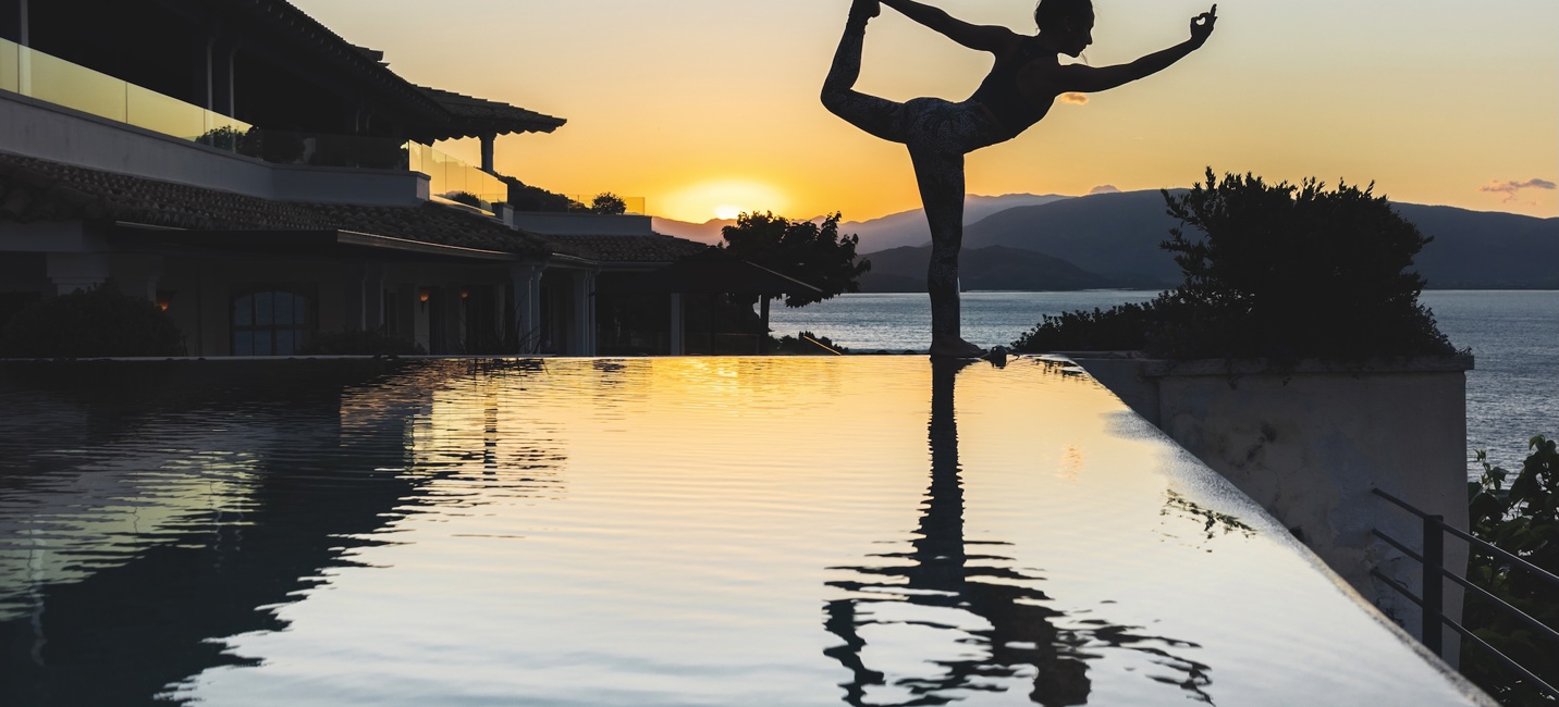 Silhouette d’une femme pratiquant le yoga au bord d’une piscine à débordement au coucher du soleil, avec les montagnes et la mer en arrière-plan.