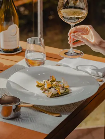 Gros plan sur une table gastronomique avec un plat raffiné d’asperges et de délicates garnitures, accompagné d’un verre de vin blanc sous la lumière naturelle du soleil.