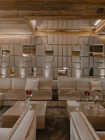 Salon luxueux avec des canapés beiges moelleux, des panneaux muraux géométriques miroitants et des accents en bois chaud, créant une atmosphère élégante et accueillante.