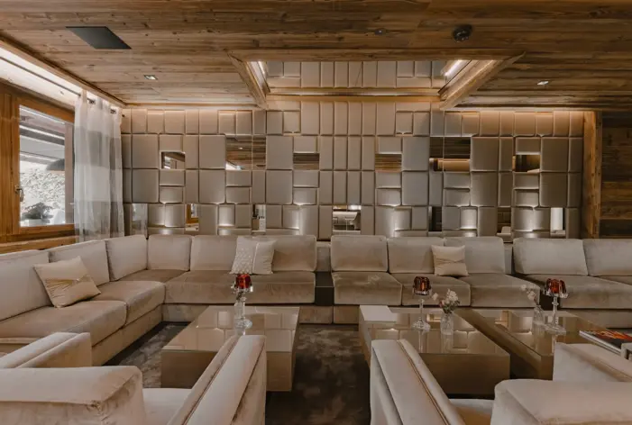 Salon luxueux avec des canapés beiges moelleux, des panneaux muraux géométriques miroitants et des accents en bois chaud, créant une atmosphère élégante et accueillante.