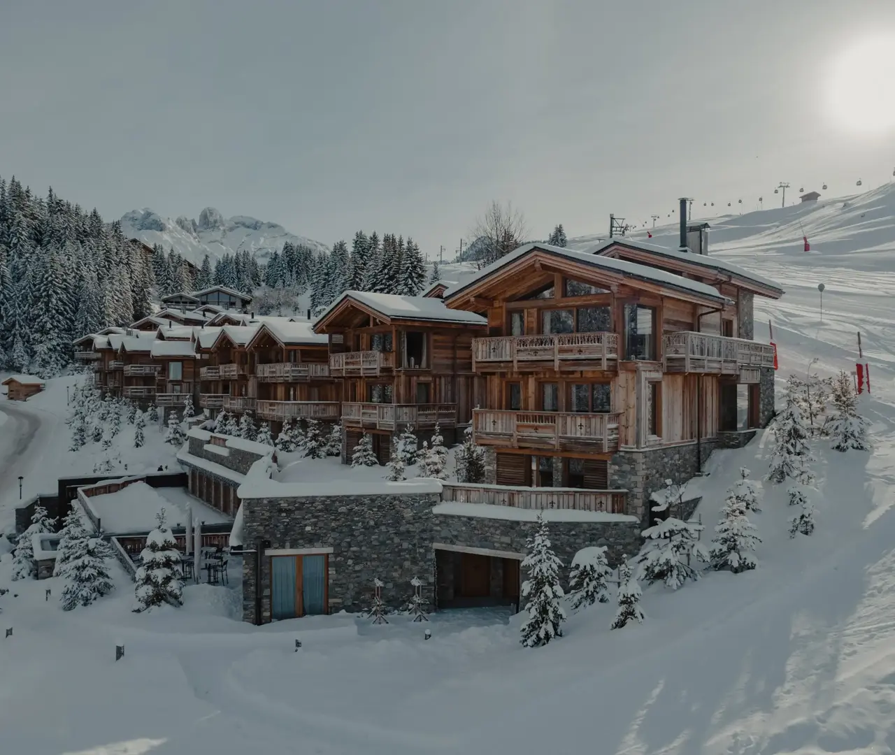 Luxurious wooden chalets nestled in a snowy alpine landscape with pine trees, ski slopes, and sunlight over the mountains.