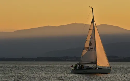 Sailboat gliding on calm water at sunset with golden light, distant mountains, and a peaceful horizon.