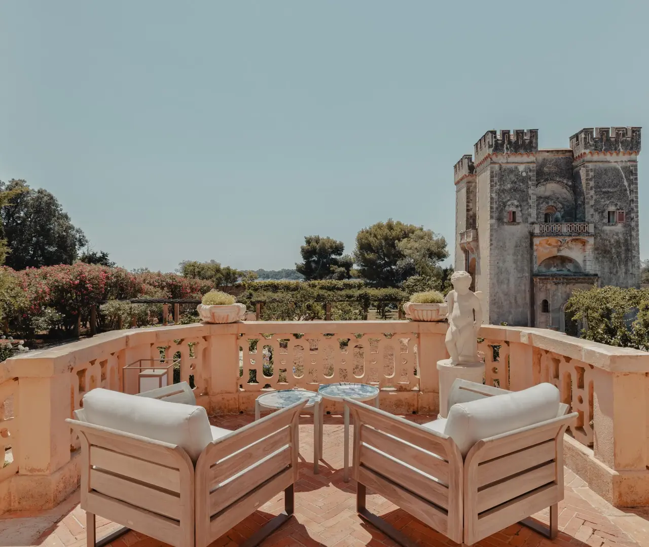 Terrasse en pierre élégante avec des fauteuils rembourrés et une petite table donnant sur des jardins et une ancienne tour en pierre sous un ciel bleu clair.