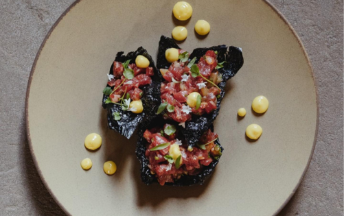 Elegant plate of finely chopped tartare on black crisps, garnished with herbs and yellow sauce dots on a beige plate.”