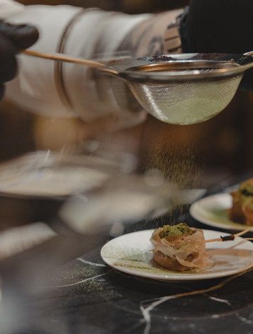 طاهٍ يحضّر مقبلات فاخرة برشّ التوابل الخضراء فوق الأطباق في مطبخ راقٍ.