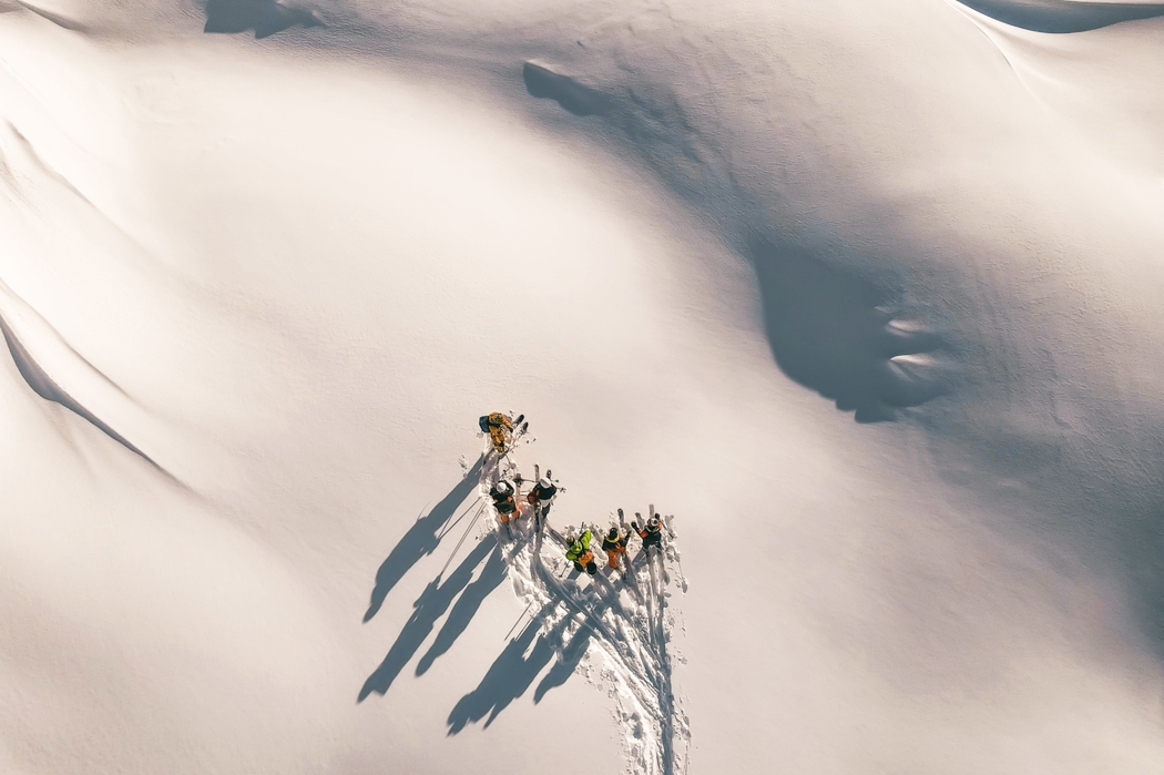 منظر جوي لمجموعة من المتزلجين يقفون على الثلج النقي، ويلقون بظلال طويلة على منحدر جبلي مضاء بأشعة الشمس.