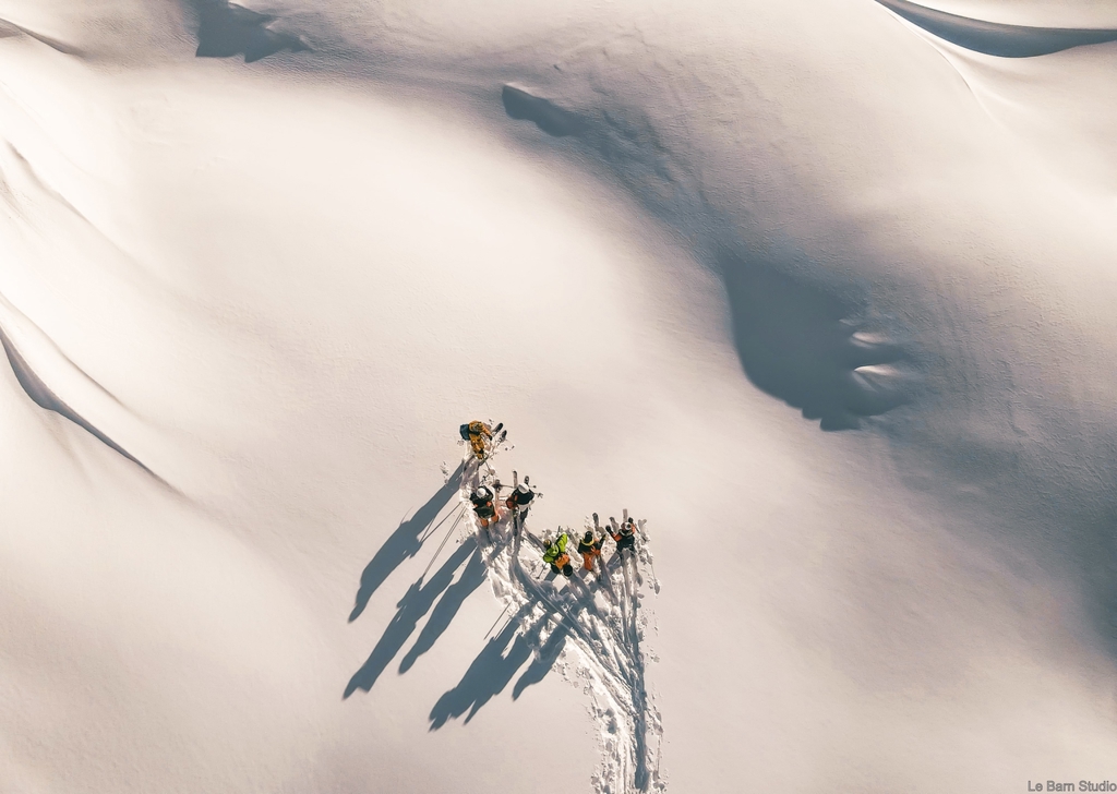 منظر جوي لمجموعة من المتزلجين يقفون على الثلج النقي، ويلقون بظلال طويلة على منحدر جبلي مضاء بأشعة الشمس.