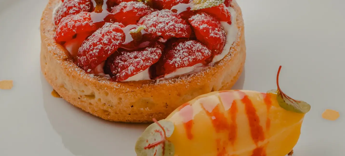 A delicate raspberry tart dusted with sugar sits beside a smooth scoop of mango sorbet drizzled with red coulis on a pristine white plate.