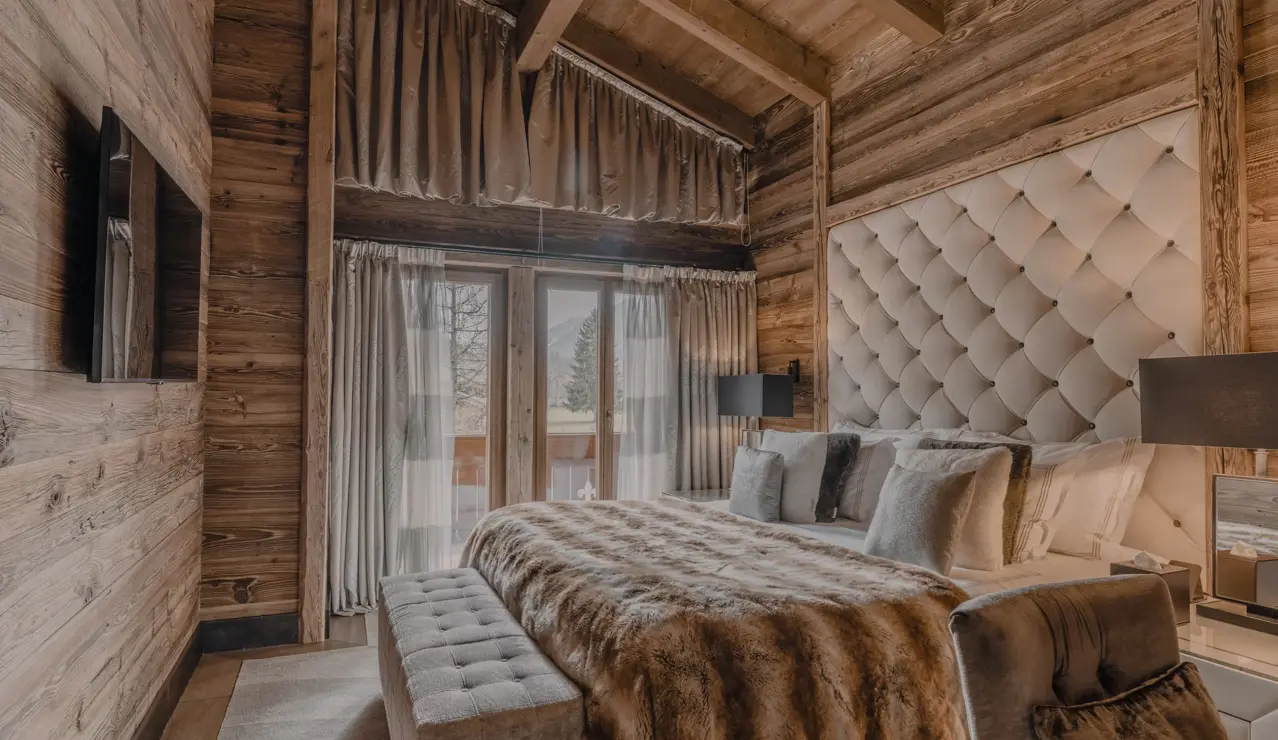 Luxurious alpine bedroom with wooden walls and exposed beams, featuring a tufted headboard, fur throw, and soft lighting that creates a warm, inviting mountain retreat atmosphere.