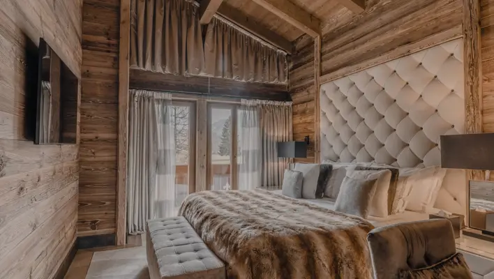 Luxurious alpine bedroom with wooden walls and exposed beams, featuring a tufted headboard, fur throw, and soft lighting that creates a warm, inviting mountain retreat atmosphere.