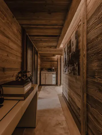 Warmly lit chalet hallway with wooden walls and ceiling, decorated with books, artwork, and elegant accessories, creating a cozy and refined alpine ambiance.